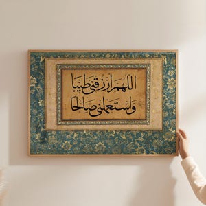 Op de afbeelding: Een ingelijst drukwerk met een decoratieve rand van teal en gouden bloemmotieven. De print bevat een kalligrafisch opschrift in zwarte inkt op een beige achtergrond. Het opschrift luidt: "Allah is de voorziener van alle dingen, en Hij is de meest barmhartige van de barmhartigen."