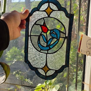 May include: Stained glass panel with a floral design. The panel features a red tulip with blue leaves and stems, set against a textured, clear background. The border is blue with yellow accents. A small metal loop is attached for hanging.