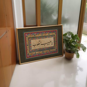 May include: A framed piece of Islamic calligraphy in black ink on a cream background. The calligraphy is surrounded by a colorful floral border. The frame is gold with a green mat.