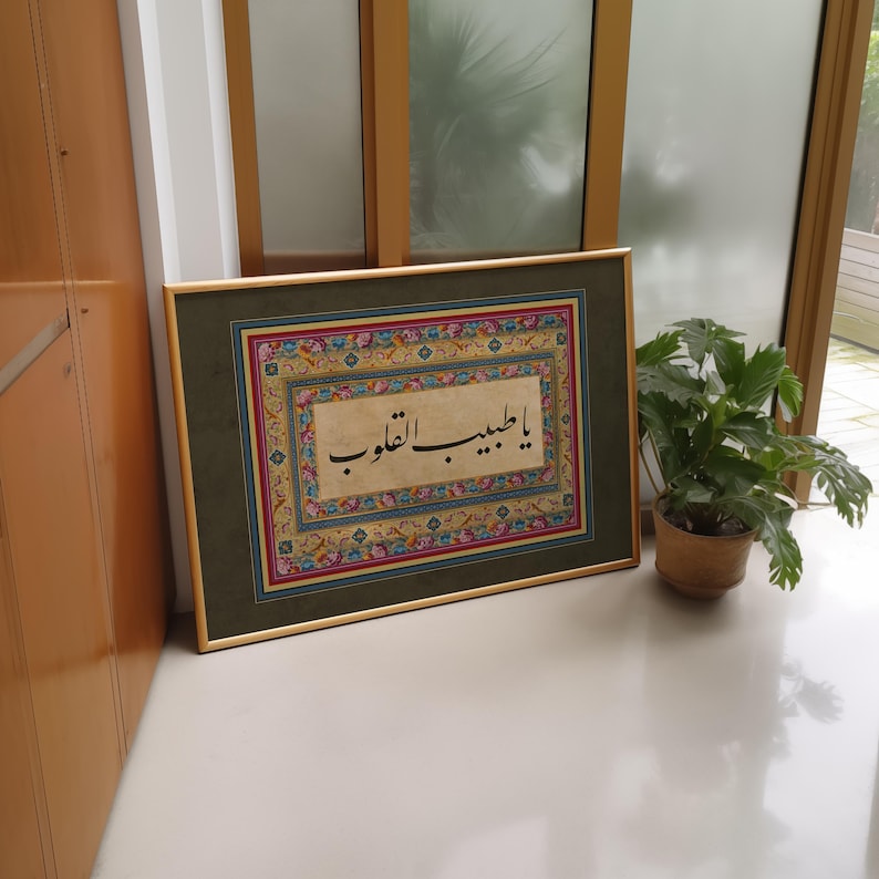 May include: A framed piece of Islamic calligraphy in black ink on a cream background. The calligraphy is surrounded by a colorful floral border. The frame is gold with a green mat.