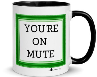 Puede incluir: Taza de café de cerámica blanca con borde y asa negros. La taza tiene un borde verde alrededor de un rectángulo blanco con el texto "YOU'RE ON MUTE" en negro.
