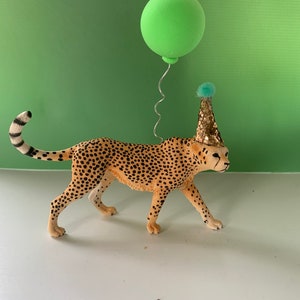 May include: A cheetah figurine with black spots on a tan body, wearing a gold glitter party hat with a small blue pom-pom on top. A thin silver wire extends from the back of the cheetah, holding a round, matte light green balloon above it.