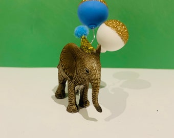 Può includere: Una piccola statuina di elefante marrone che indossa un cappello da festa e tiene in mano due palloncini, uno blu e uno bianco con glitter dorati.