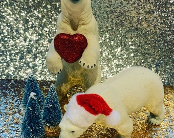 May include: Two white polar bear figurines, one holding a red heart, the other wearing a red and white Santa hat, standing on a gold glitter surface with two small blue pine trees.