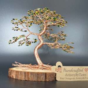 May include: Handcrafted copper wire tree sculpture with green leaves on a wooden base. The tree is styled like a bonsai and has a tag that reads "Handcrafted ArtistryByTawn Etsy/Instagram: ArtistryByTawn".