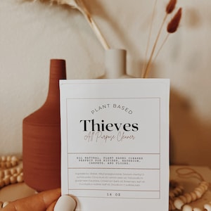 May include: A white label for "Thieves All Purpose Cleaner" with black text. The label states it is plant-based and all-natural, perfect for kitchen, bathroom, carpets, and floors. The label is 16 oz. and held in front of a terracotta vase.