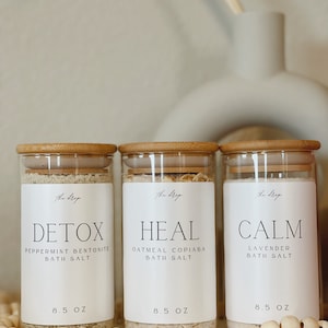 May include: Three glass jars with bamboo lids filled with bath salts. The labels read "DETOX Peppermint Bentonite Bath Salt 8.5 oz", "HEAL Oatmeal Copaiba Bath Salt 8.5 oz", and "CALM Lavender Bath Salt 8.5 oz".