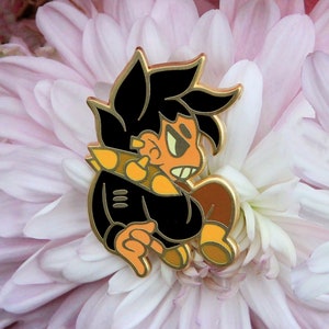 May include: Gold-colored enamel pin featuring a cartoon character with black hair, a black jacket, and orange accents. The character has a serious expression and is wearing a black jacket with a white stripe.