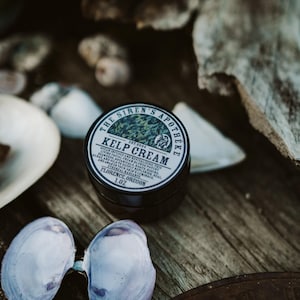 Może przedstawiać: Mały czarny słoiczek z kremem z wodorost&oacute;w z białą etykietą, na kt&oacute;rej widnieje napis "The Siren's Apotheke Est. 2024 Kelp Cream Ocean Infused and Revitalizing This Living Facial Cream Blends Potent Algae and Oils to Deeply Replenish Real Seaweeds Botanicals Roots Res and Awaken You'r Skin to Ethereal Radiance Florence, Oregon 1.0z"