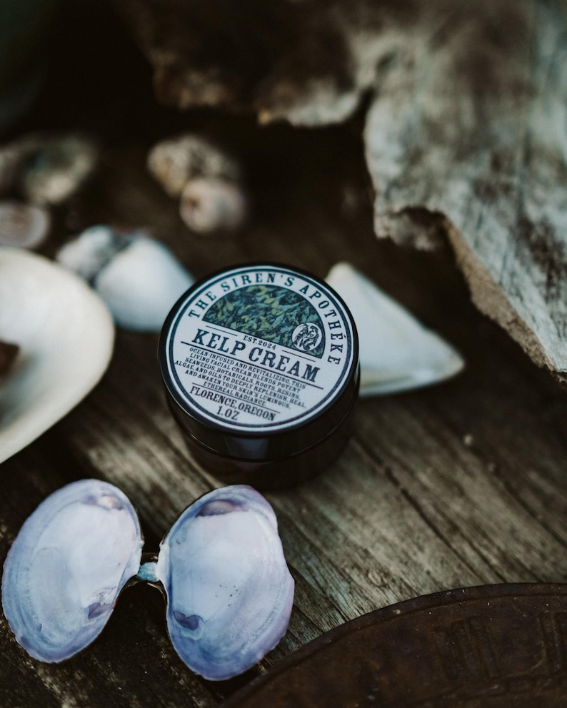 Może przedstawiać: Mały czarny słoiczek z kremem z wodorost&oacute;w z białą etykietą, na kt&oacute;rej widnieje napis "The Siren's Apotheke Est. 2024 Kelp Cream Ocean Infused and Revitalizing This Living Facial Cream Blends Potent Algae and Oils to Deeply Replenish Real Seaweeds Botanicals Roots Res and Awaken You'r Skin to Ethereal Radiance Florence, Oregon 1.0z"
