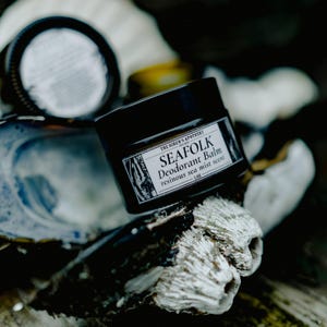 May include: A black jar of "SEAFOLK Deodorant Balm" with a white label, resting on a textured, white and grey surface. The label indicates a "resinous sea mist scent" and the jar contains 28g of product. Another jar is visible in the background.