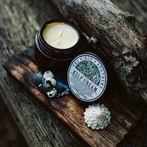 Może przedstawiać: Mały słoiczek z jasnoż&oacute;łtym kremem z wodorost&oacute;w z czarną i białą etykietą z napisem "The Siren's Apotheke Est 2024 Kelp Cream". Słoiczek stoi na kawałku drewna unoszącego się na wodzie z muszelkami.