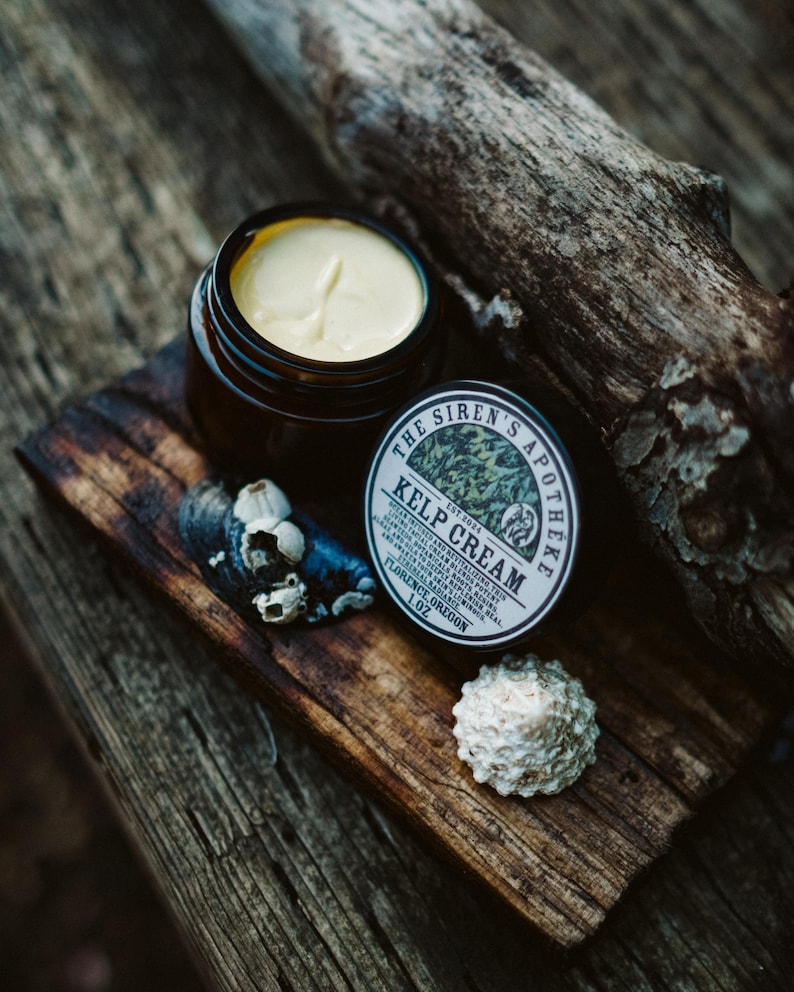 Może przedstawiać: Mały słoiczek z jasnoż&oacute;łtym kremem z wodorost&oacute;w z czarną i białą etykietą z napisem "The Siren's Apotheke Est 2024 Kelp Cream". Słoiczek stoi na kawałku drewna unoszącego się na wodzie z muszelkami.