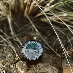 Może przedstawiać: Mały czarny słoik z białą etykietą, na kt&oacute;rej widnieje napis "The Siren's Apotheka Est 2024 Kelp Cream Ocean infused and revitalizing this living facial cream utilizes potent algae and botanicals to deeply replenish and revitalize skin.  Awaken your natural radiance.  Florence, Oregon 1.0 oz."