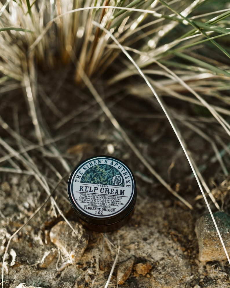 Może przedstawiać: Mały czarny słoik z białą etykietą, na kt&oacute;rej widnieje napis "The Siren's Apotheka Est 2024 Kelp Cream Ocean infused and revitalizing this living facial cream utilizes potent algae and botanicals to deeply replenish and revitalize skin.  Awaken your natural radiance.  Florence, Oregon 1.0 oz."