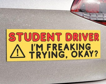 May include: Yellow rectangular bumper sticker with the text "STUDENT DRIVER I'M FREAKING TRYING, OKAY?" in red and black lettering. A black triangle with an exclamation point is on the left side. The sticker is affixed to the back of a silver car.