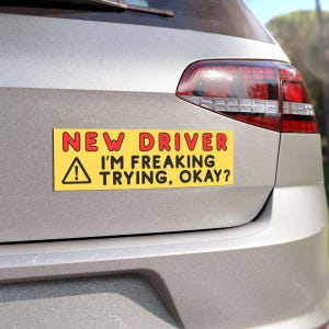 Peut inclure: Un autocollant de pare-chocs rectangulaire jaune avec le texte "NEW DRIVER" en lettres rouges au-dessus de "I'M FREAKING TRYING, OKAY?" en noir. Un triangle noir avec un point d'exclamation est à gauche.