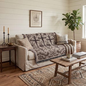 a cozy living room with a beige sofa adorned with a patterned throw blanket, a wooden coffee table, and a framed picture on the wall. The room is decorated with a potted plant, a candle holder, and a book, creating a warm and inviting atmosphere.