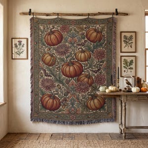 May include: A rectangular woven wall hanging featuring a repeating botanical pattern of orange pumpkins, muted pink chrysanthemums, and green foliage. The textile has a fringed bottom edge and is suspended from a wooden branch with metal clips.