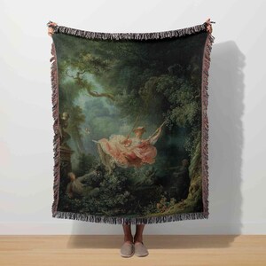 May include: Throw blanket featuring a classical painting of a woman on a swing in a lush, green forest. The artwork depicts a romantic scene with soft pink and white clothing, surrounded by trees and foliage. The blanket has a fringed edge.