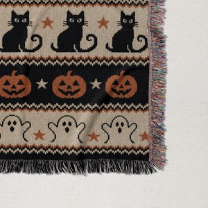 Knitted throw blanket with black cats, pumpkins, and ghosts on beige background.