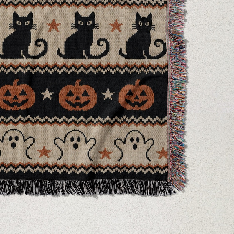 Knitted throw blanket with black cats, pumpkins, and ghosts on beige background.