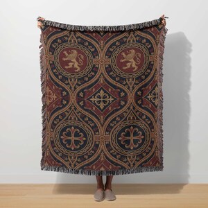 May include: A woven throw blanket with a repeating pattern of medallions, crosses, and fleur-de-lis in navy blue, burgundy, and gold. The design features heraldic lions and intricate geometric shapes. The blanket has a fringed edge.