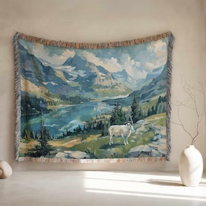 May include: Woven tapestry depicting a mountain landscape with a lake, trees, and a white goat. The artwork uses shades of blue, green, and beige, with a fringed border. The tapestry is displayed on a wall.