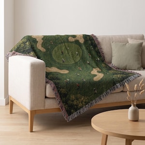 May include: A woven throw blanket featuring an aerial view of a golf course with green fairways, sand bunkers, and small figures of golfers in various positions. The blanket has a fringed edge and a dark green, tan, and brown color palette.