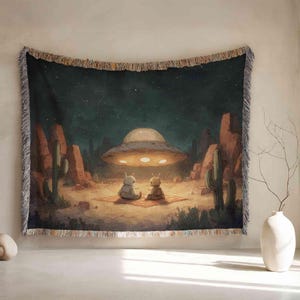 May include: Woven tapestry featuring a desert scene with a UFO hovering over two small robot-like figures. The artwork includes cacti, red rock formations, and a starry night sky. The tapestry has a fringed edge.
