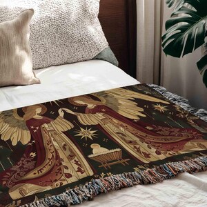 a bed with a decorative blanket on it, which has a detailed and intricate design with angels, a nativity scene, and stars. The blanket is placed on a white bedspread, and there is a window with a plant visible in the background.