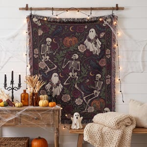 May include: A woven tapestry blanket featuring a dark purple background with illustrations of skeletons, ghosts, pumpkins, roses, and ravens. The rectangular textile has a fringed edge and is displayed hanging from a wooden rod with string lights.