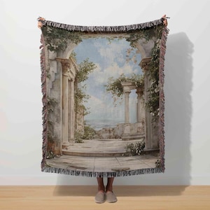 May include: A woven throw blanket featuring a classical architectural scene with columns, arches, and a sky with clouds. The design incorporates muted tones of beige, blue, and green, with a fringed edge.
