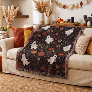 May include: A woven throw blanket featuring a dark background with a repeating pattern of white ghosts, orange pumpkins, brown acorns, and autumn leaves. The rectangular blanket has fringed edges and a detailed botanical design with swirling vines.