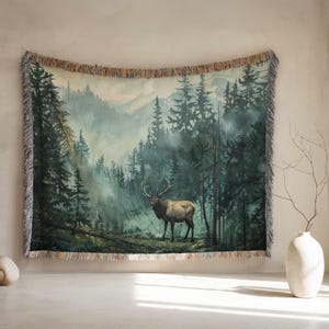 May include: A woven tapestry depicting an elk in a misty forest. The artwork shows a detailed landscape with tall evergreen trees and a mountainous backdrop. The tapestry has a fringed edge and is designed for wall hanging.