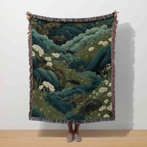 May include: A woven throw blanket featuring a landscape design with a dark teal and green color palette. The design includes white flowers, rocks, and foliage. The blanket has a fringed edge.