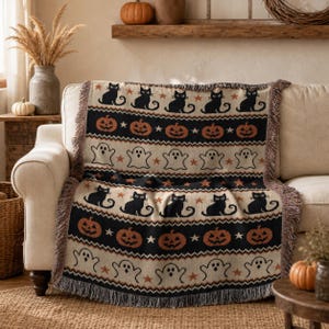 a cozy living room with a white sofa and a decorative Halloween-themed throw blanket draped over it. The blanket has a repeating pattern of black cats, jack-o-lanterns, and ghostly figures, creating a festive and spooky atmosphere.