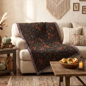 May include: A woven throw blanket featuring a dark, intricate pattern of orange and green autumn leaves, spiderwebs, and bats. The blanket has a fringed edge and is draped over a light-colored sofa, showcasing a detailed botanical and seasonal motif.