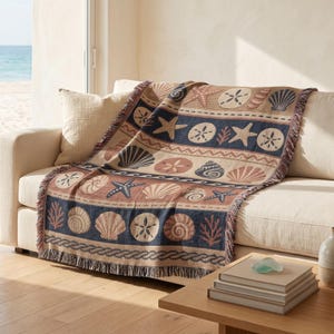 May include: A woven throw blanket featuring a repeating pattern of seashells, starfish, and sand dollars in shades of navy blue, beige, and terracotta. The blanket has a fringed edge and is draped over a light-coloured sofa in a bright room.
