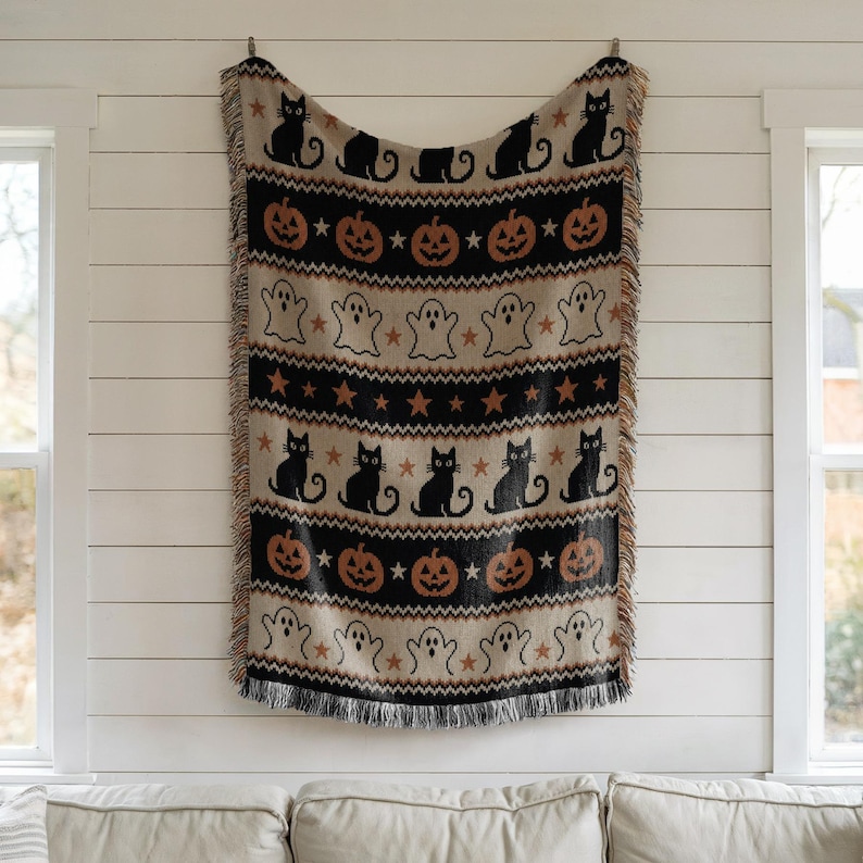 Woven Halloween blanket with black cats, pumpkins, and ghosts on a cream background.
