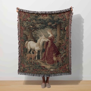 May include: A tapestry throw blanket depicting a medieval scene with a woman, a unicorn, and deer in a forest. The blanket has a detailed design with a fringed edge and measures approximately 152 cm by 127 cm.
