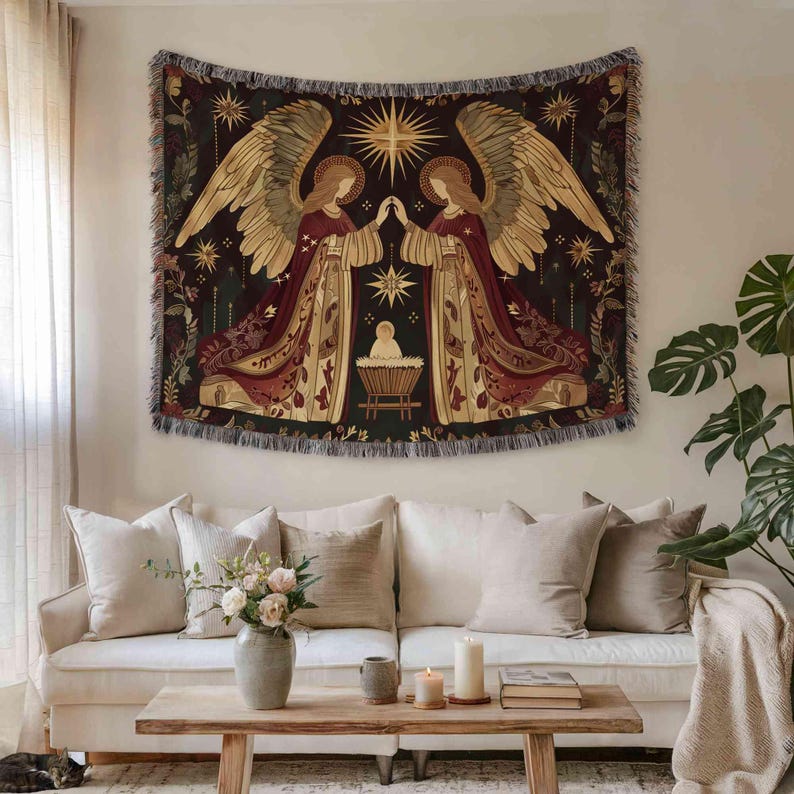 a living room with a large tapestry hanging on the wall above a white sofa. The tapestry depicts two angels with wings, one on each side, standing in front of a baby in a crib. The room also includes a coffee table with a vase of flowers, a book, and a candle.