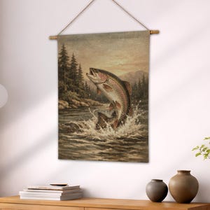 Op de afbeelding: Een wandkleed met een vintage illustratie van een regenboogforel die uit het water springt. Het kunstwerk heeft een natuurlijk kleurenpalet met een bos- en bergachtergrond. De print is opgehangen aan een houten roede met een natuurlijk koord.