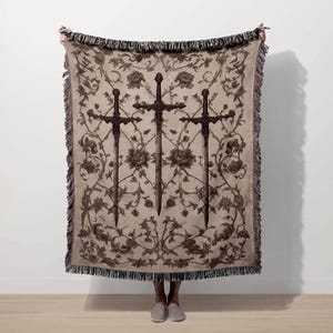A person is holding up a large blanket with a floral design and three crossed swords on it.