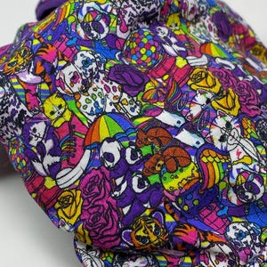 K&ouml;nnte beinhalten: Ein farbenfroher Stoff mit einem sich wiederholenden Muster aus Totenk&ouml;pfen, Rosen und anderen skurrilen Illustrationen in Lila, Rosa, Gelb und Blau. Der Stoff ist gefaltet und zeigt das detaillierte Design.