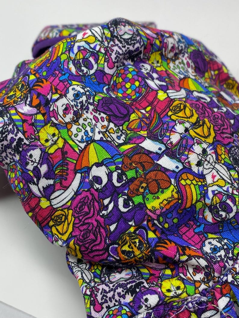 K&ouml;nnte beinhalten: Ein farbenfroher Stoff mit einem sich wiederholenden Muster aus Totenk&ouml;pfen, Rosen und anderen skurrilen Illustrationen in Lila, Rosa, Gelb und Blau. Der Stoff ist gefaltet und zeigt das detaillierte Design.