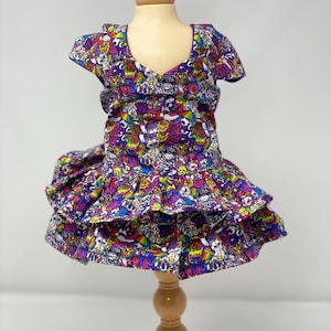 K&ouml;nnte beinhalten: Ein leuchtend lila Kleid mit V-Ausschnitt und kurzen &Auml;rmeln, pr&auml;sentiert auf einer Holzpuppe. Das Kleid hat ein farbenfrohes, sich wiederholendes Muster aus Cartoonfiguren und Totenk&ouml;pfen. Der Rock hat ger&uuml;schte Schichten.