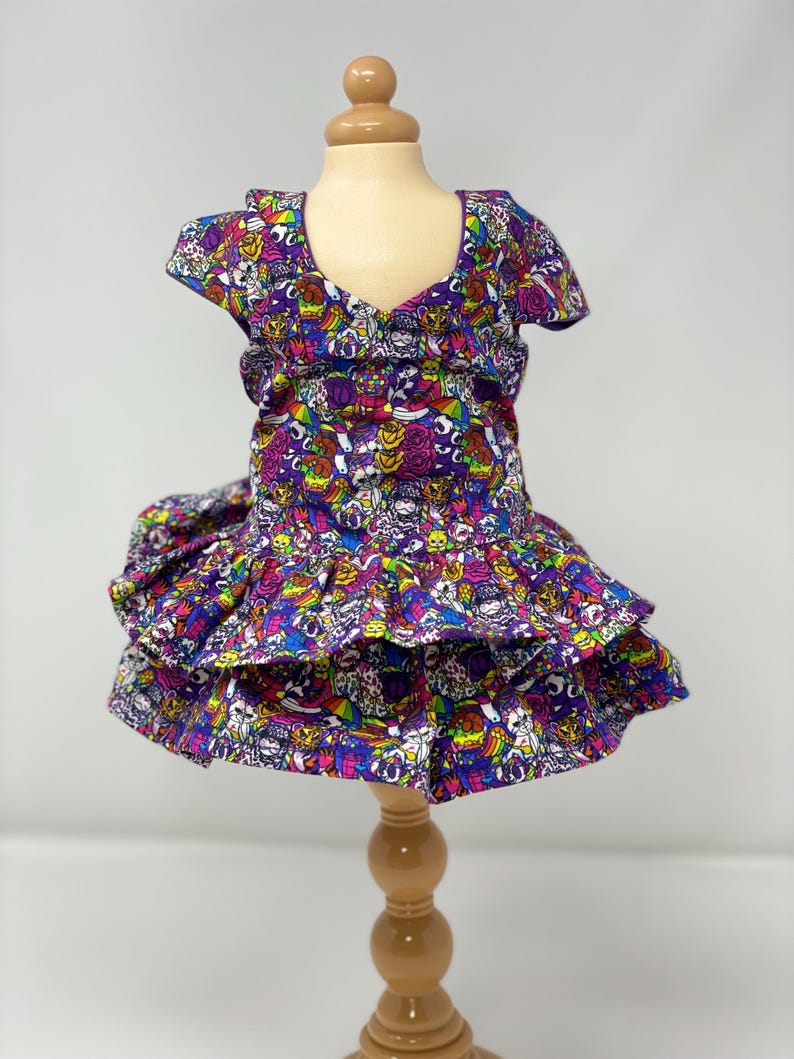 K&ouml;nnte beinhalten: Ein leuchtend lila Kleid mit V-Ausschnitt und kurzen &Auml;rmeln, pr&auml;sentiert auf einer Holzpuppe. Das Kleid hat ein farbenfrohes, sich wiederholendes Muster aus Cartoonfiguren und Totenk&ouml;pfen. Der Rock hat ger&uuml;schte Schichten.