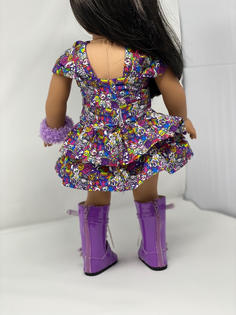 K&ouml;nnte beinhalten: Eine Puppe in einem lila Kleid mit buntem Totenkopfmuster, einem passenden lila flauschigen Armband und gl&auml;nzenden lila Stiefeln. Das Kleid hat einen ger&uuml;schten Rock und kurze &Auml;rmel. Die Stiefel haben Schn&uuml;rsenkel und Rei&szlig;verschl&uuml;sse.