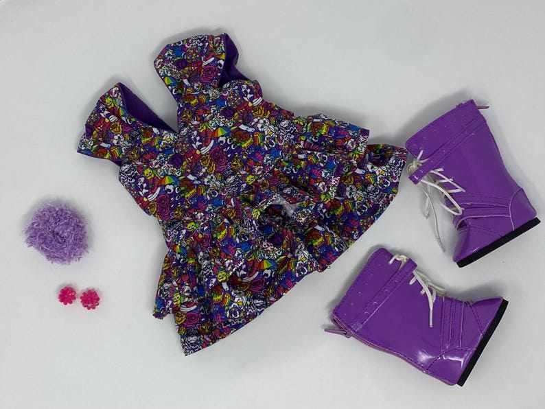 K&ouml;nnte beinhalten: Ein Puppen-Outfit mit einem farbenfrohen gemusterten Kleid, lila Stiefeln mit wei&szlig;en Schn&uuml;rsenkeln, einem lila Pompon und rosa bl&uuml;tenf&ouml;rmigen Ohrringen. Das Kleid hat einen lebendigen Druck mit verschiedenen Formen und Farben.